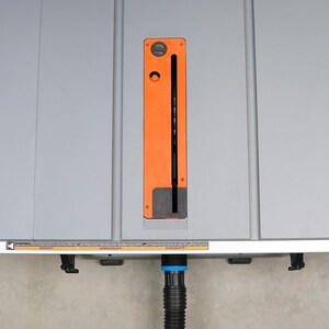 May include: A gray table saw with an orange blade guard and a black power cord. The saw has a dust collection port on the front.