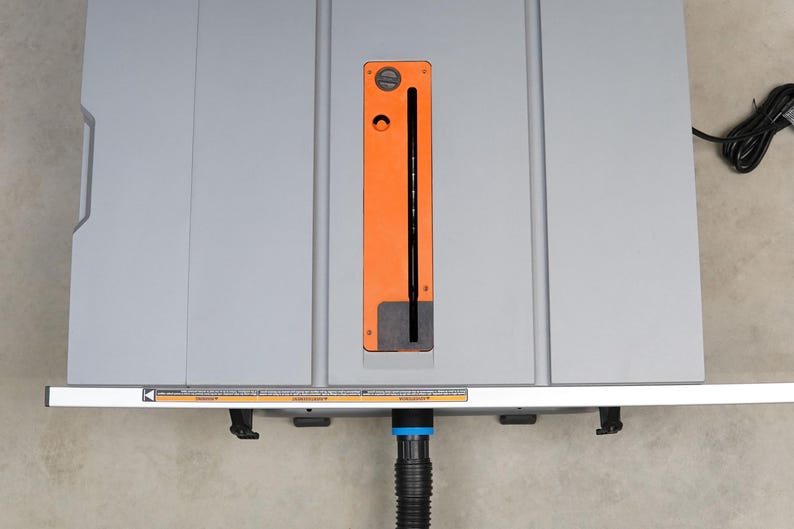 May include: A gray table saw with an orange blade guard and a black power cord. The saw has a dust collection port on the front.