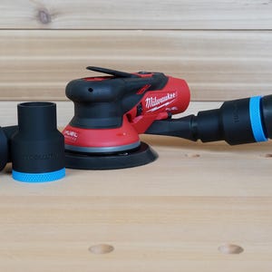 Puede incluir: Una lijadora orbital Milwaukee FUEL roja y negra con un accesorio de recolección de polvo negro y dos adaptadores negros con detalles azules. La lijadora está sobre una superficie de madera clara.