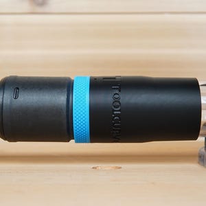 May include: Black and blue tool accessory with the text "TOOLCURVE" on the side. The accessory is connected to a black and green hose and a clear plastic component. The background is a light brown wooden surface.