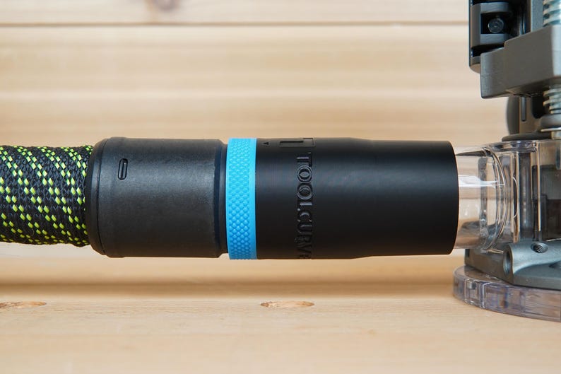 May include: Black and blue tool accessory with the text "TOOLCURVE" on the side. The accessory is connected to a black and green hose and a clear plastic component. The background is a light brown wooden surface.