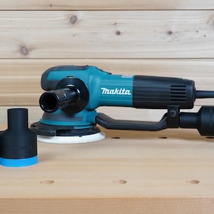 Puede incluir: Una lijadora orbital aleatoria Makita turquesa y negra con un mango y cable de alimentación negros. Dos accesorios negros de recogida de polvo con detalles azules están delante de la lijadora. La lijadora está sobre una superficie de madera.