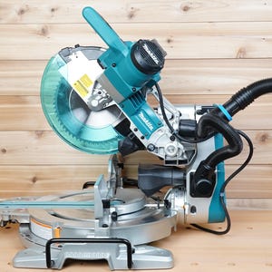 May include: A teal and silver miter saw with a clear blade guard and a black dust collection hose. The saw has a silver base and a teal handle. The Makita brand name is visible on the saw.