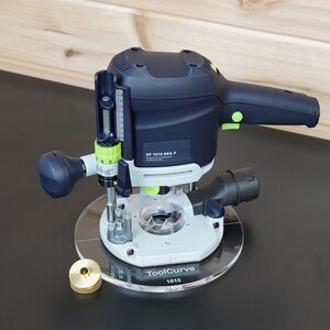 Peut inclure: Une défonceuse électrique bleu foncé et gris clair avec des accents verts, dotée d'une plaque de base circulaire transparente marquée ToolCurve 1010. L'appareil comprend une échelle de réglage de profondeur, une poignée et une pièce circulaire en laiton.