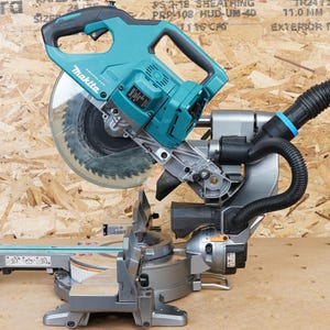 May include: A teal and silver Makita power mitre saw with a dust collection hose attached. The saw is on a wooden surface with a sheet of plywood behind it.