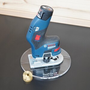 Könnte beinhalten: Ein blauer und schwarzer Bosch 12V Akku-Kantenfräser montiert auf einer runden, transparenten Acrylplatte mit dem Aufdruck ToolCurve BOSCH 12V TRIM, ergänzt durch eine kleine messingfarbene Metall-Führungshülse neben der Basis.