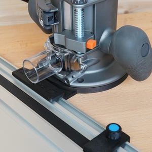 May include: A gray and black router with a clear plastic dust collection tube is mounted on a wooden surface. The router has a black handle and an orange button. A silver guide rail with the Milwaukee logo is visible.