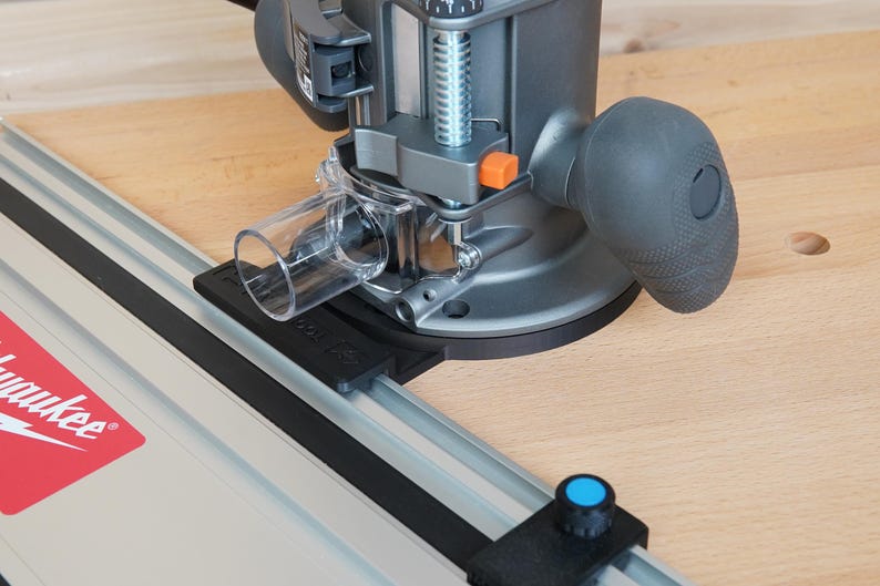May include: A gray and black router with a clear plastic dust collection tube is mounted on a wooden surface. The router has a black handle and an orange button. A silver guide rail with the Milwaukee logo is visible.