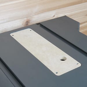 May include: A close-up shot of a dark gray table saw with a light-colored rectangular insert. The insert has a circular hole and is secured with four screws. The background is a light-colored wooden surface.