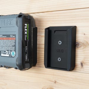 May include: A black and gray FLEX 24V lithium-ion battery pack and a black battery holder mounted on a wooden surface. The battery pack has a green and white label with specifications. The holder is rectangular with the FLEX logo.