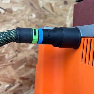 May include: A black and green dust collection hose with a black connector attached to an orange dust collector. The connector has a green and blue label that reads "Festool SYS-I".