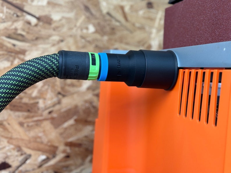 May include: A black and green dust collection hose with a black connector attached to an orange dust collector. The connector has a green and blue label that reads "Festool SYS-I".