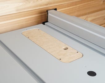 May include: A close-up of a gray table saw surface with a light brown insert. The saw has a silver rail and a black button. The background is a light brown wooden wall.