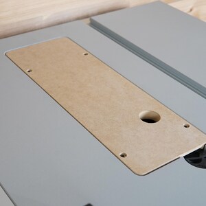 May include: A close-up shot of a gray table saw surface with a rectangular, light brown insert. The insert has a circular hole and several smaller holes. The saw's surface is light gray, and the background is a light wood.