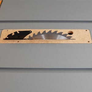 Puede incluir: Primer plano de una sierra de mesa con una hoja plateada y un protector de hoja negro. La hoja es parcialmente visible a trav&eacute;s de una abertura de madera en la sierra de mesa.