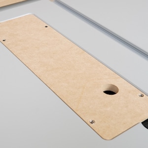 May include: A light brown rectangular wooden insert with a circular hole and three screw holes, set into a light gray surface. The insert is part of a larger piece of equipment.