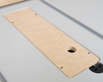 May include: A light brown rectangular wooden insert with a circular hole and three screw holes, set into a light grey surface. The insert is part of a larger piece of equipment.