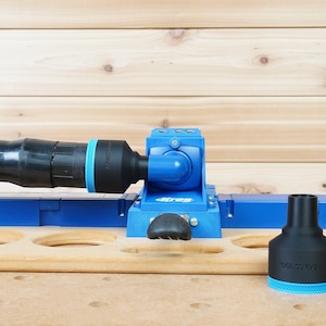 Könnte beinhalten: Eine blaue Kreg-Taschenlochlehre mit einem schwarzen und orangefarbenen Schlauch. Ein schwarzer Adapter mit dem Text "TOOLCURVE" befindet sich auf der rechten Seite. Die Vorrichtung befindet sich auf einer Holzoberfläche mit mehreren Löchern.