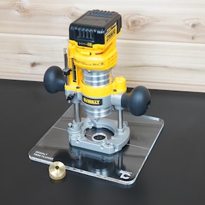 May include: A yellow and black DeWalt cordless trim plunge router mounted on a clear acrylic base plate. The tool features two black side handles, a metal depth adjustment rod, and a small brass circular guide component resting on the base.