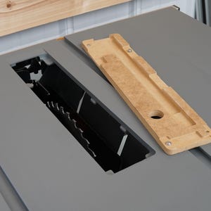Puede incluir: Primer plano de una sierra de mesa con un inserto de madera. El inserto es de color marr&oacute;n claro y tiene una ranura cortada. La sierra de mesa es gris y tiene una cuchilla negra.