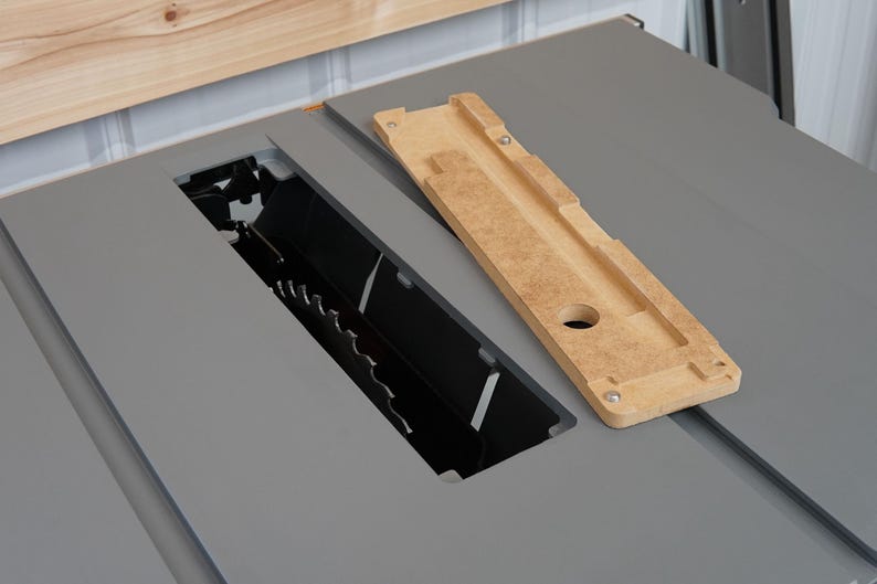 Puede incluir: Primer plano de una sierra de mesa con un inserto de madera. El inserto es de color marr&oacute;n claro y tiene una ranura cortada. La sierra de mesa es gris y tiene una cuchilla negra.