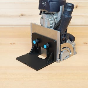 Op de afbeelding: Een lamellenfrees met een zwarte kunststof voetplaat met twee blauwe instelknoppen, een zilverkleurige metalen geleider en een zwarte stofafzuigpoort. De motorbehuizing is donkerblauw en zilverkleurig met zichtbare schroeven en onderdelen.