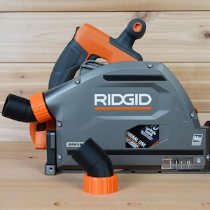 May include: A gray and orange RIDGID circular saw with a dust port and a 7-1/4 inch blade. The saw has a black dust collection hose attached to the dust port. The saw is sitting on a wooden surface.