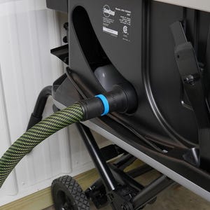 Puede incluir: Una sierra de mesa negra y gris con un puerto de recolección de polvo. Una manguera verde y negra está conectada al puerto. La sierra tiene una base de metal negro con ruedas y un brazo de metal negro.