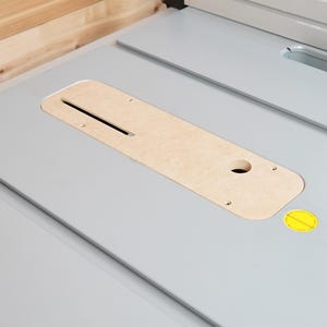 May include: A light gray table saw surface with a wooden insert. The insert has a long slot and a round hole. A yellow sticker is visible on the surface. The background is a light wood.