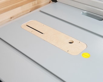 May include: A light grey table saw surface with a wooden insert. The insert has a long slot and a round hole. A yellow sticker is visible on the surface. The background is light wood.