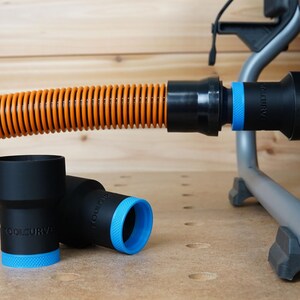 Op de afbeelding: Een close-up van een zwarte en oranje stofzuigerslang met een zwarte stofzuigeradapter. Twee zwarte stofzuigeradapters met blauwe ringen liggen op een houten oppervlak.