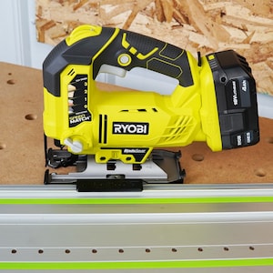 May include: A yellow and black Ryobi jigsaw with a black battery pack. The tool has a handle and a blade guard. The jigsaw is on a brown surface with a green and silver guide rail.