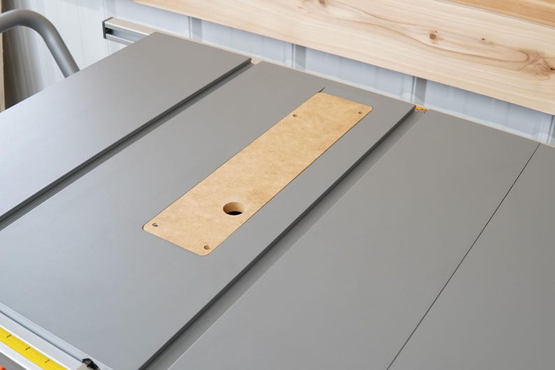 Puede incluir: Una sierra de mesa gris con un panel de inserci&oacute;n de madera. El panel tiene un agujero en el centro y est&aacute; rodeado por una superficie gris.