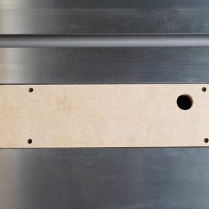 May include: A close-up shot of a table saw insert. The insert is a light tan color, rectangular with rounded ends, and has a circular hole. It is set into a silver-colored metal surface and secured with screws.