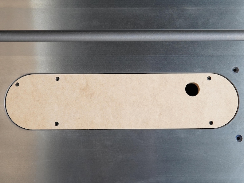 May include: A close-up shot of a table saw insert. The insert is a light tan color, rectangular with rounded ends, and has a circular hole. It is set into a silver-colored metal surface and secured with screws.