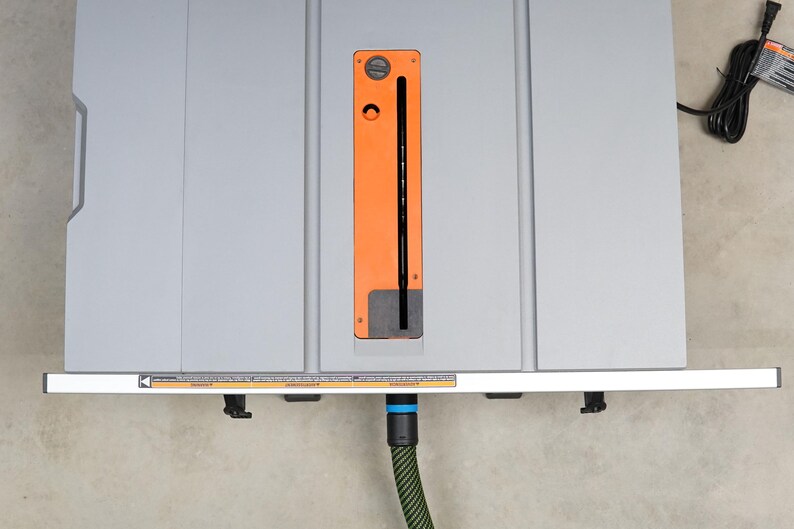 May include: A grey table saw with an orange and black blade guard. The saw has a dust collection port on the side and a power cord plugged in. The saw is sitting on a concrete floor.