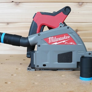 Op de afbeelding: Een grijze en rode Milwaukee FUEL cirkelzaag met een zwarte stofafvoer en een zwarte en blauwe stofafzuigadapter. De zaag staat op een houten oppervlak.