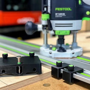 以下が含まれることがあります： 緑色のノブと銀色のネジが付いた黒いルーター治具。治具は、Festool OF 1400 EBルーターの隣の木製表面の上に置かれています。ルーターは黒と緑で、銀色のベースが付いています。