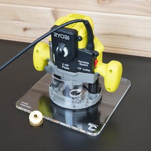 Könnte beinhalten: Eine gelb-schwarze Ryobi Oberfräse mit variabler Drehzahlregelung, 2 PS Motor und 1/2 Zoll Spannzange, montiert auf einer quadratischen, transparenten Acryl-Grundplatte, mit einer kleinen Messing-Führungshülse daneben platziert.