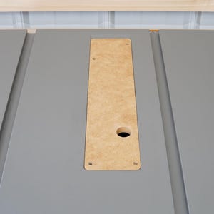 Puede incluir: Primer plano de una superficie gris con un recorte rectangular. El recorte est&aacute; relleno con una pieza de madera marr&oacute;n claro con un agujero redondo en el centro. Hay cuatro peque&ntilde;os agujeros alrededor de los bordes de la madera.