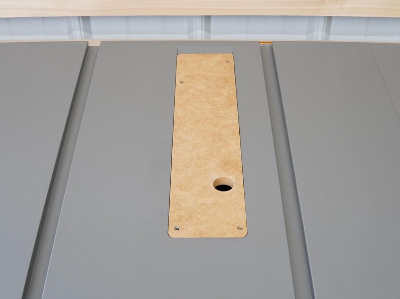 Puede incluir: Primer plano de una superficie gris con un recorte rectangular. El recorte est&aacute; relleno con una pieza de madera marr&oacute;n claro con un agujero redondo en el centro. Hay cuatro peque&ntilde;os agujeros alrededor de los bordes de la madera.
