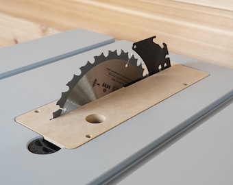 May include: A close-up of a table saw with a circular blade. The blade is silver with sharp teeth and a black protective cover. The saw is set in a light grey table with a wooden insert.