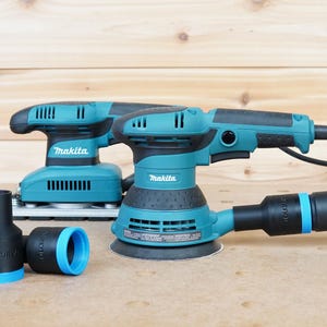 Puede incluir: Dos lijadoras eléctricas Makita azules y negras con mangueras de aspiración de polvo. Las lijadoras están sobre una superficie de madera. Una lijadora tiene una boquilla de aspiración de polvo azul y la otra tiene una boquilla de aspiración de polvo negra.