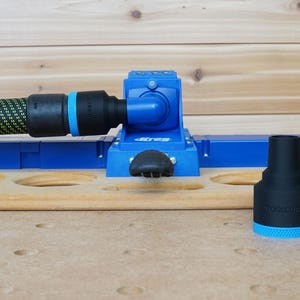Könnte beinhalten: Ein blauer und schwarzer Jig mit einem Staubabsauganschluss und einem schwarzen Knopf. Der Jig befindet sich auf einer Holzoberfläche mit zwei schwarzen und blauen Staubabsaugadaptern.