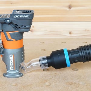 Pode incluir: Uma fresadora sem fio preta e laranja com uma porta de coleta de p&oacute; conectada a uma mangueira de coleta de p&oacute; preta com uma faixa azul. A fresadora &eacute; etiquetada como "RIDGID" e "OCTANE".