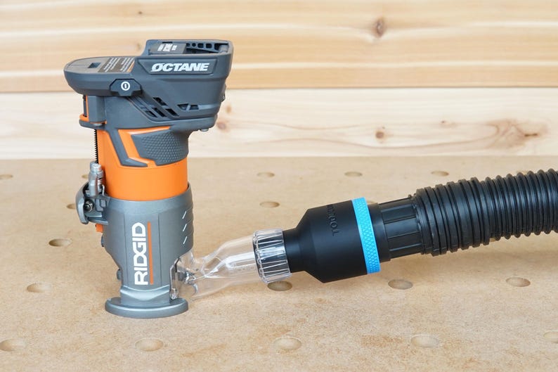 Pode incluir: Uma fresadora sem fio preta e laranja com uma porta de coleta de p&oacute; conectada a uma mangueira de coleta de p&oacute; preta com uma faixa azul. A fresadora &eacute; etiquetada como "RIDGID" e "OCTANE".