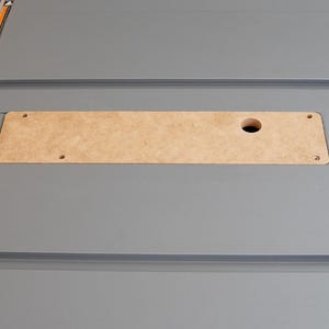 Puede incluir: Primer plano de una mesa de color gris con una inserci&oacute;n rectangular de madera. La inserci&oacute;n tiene un agujero redondo en el centro y dos peque&ntilde;os agujeros a cada lado.