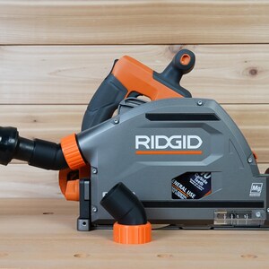 May include: A gray and orange RIDGID circular saw with a dust collection system. The saw has a black dust port and a clear dust collection container. The saw is on a wooden surface.