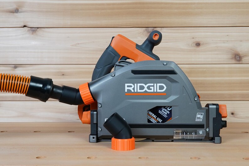 May include: A gray and orange RIDGID circular saw with a dust collection system. The saw has a black dust port and a clear dust collection container. The saw is on a wooden surface.