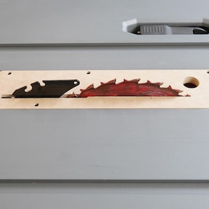 May include: A close-up of a table saw blade. The blade is red and has sharp teeth. It is set in a light brown insert. The table is gray and has a black handle.